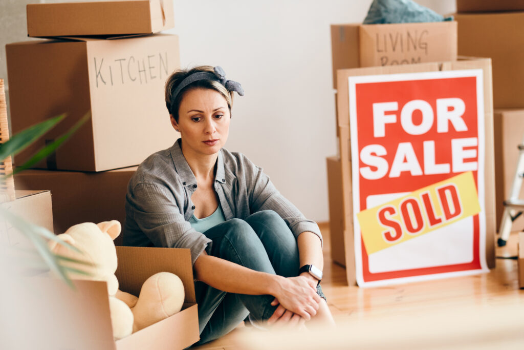 Pensive woman sitting on the floor among cardboard boxes and feeling sad after selling hew apartment.